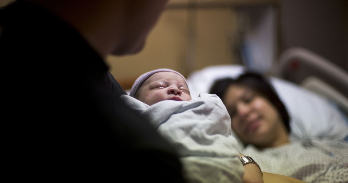 szuletes - Gyerekszoba Újszülött baba apja karjaiban a szülőszobán, háttérben az édesanyja