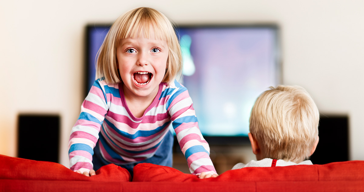 Kislány kiált a kanapén állva, testvére a tévét nézi