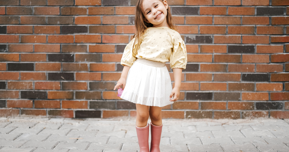 Kislány fehér ruhában és rózsaszín gumicsizmában mosolyog téglafal előtt
