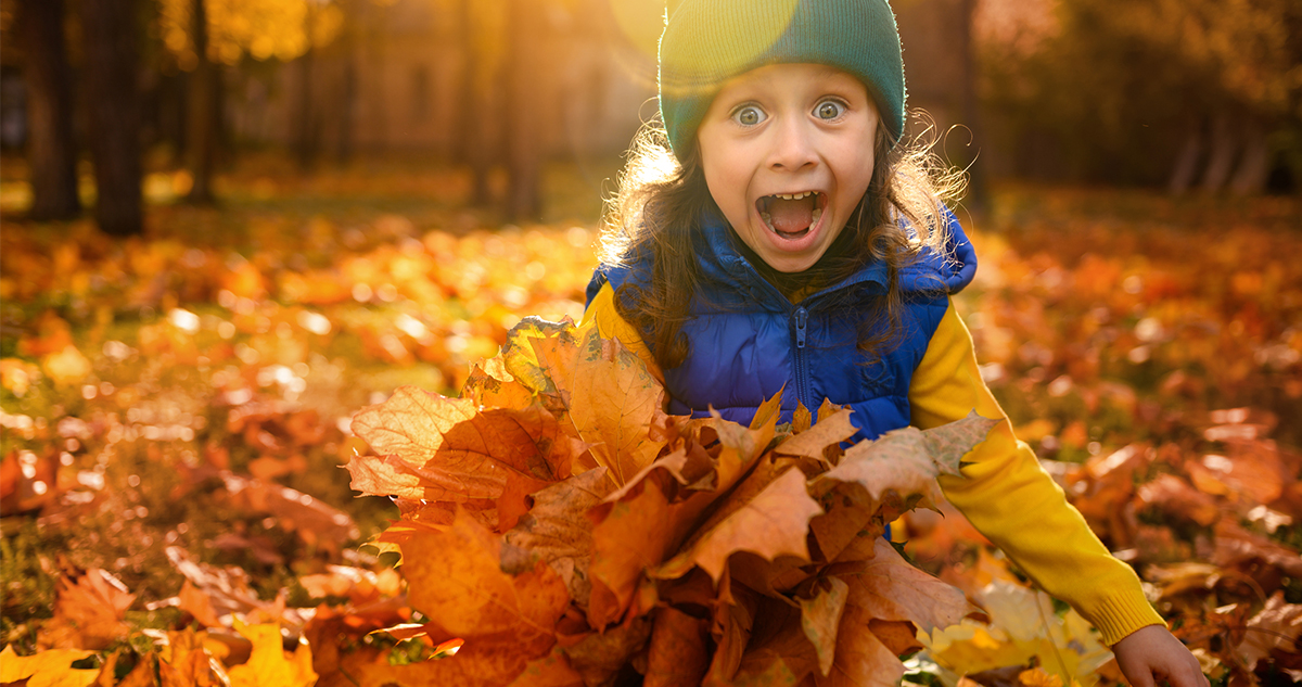 Vidám kislány őszi levelek között, napsütésben, sapkában