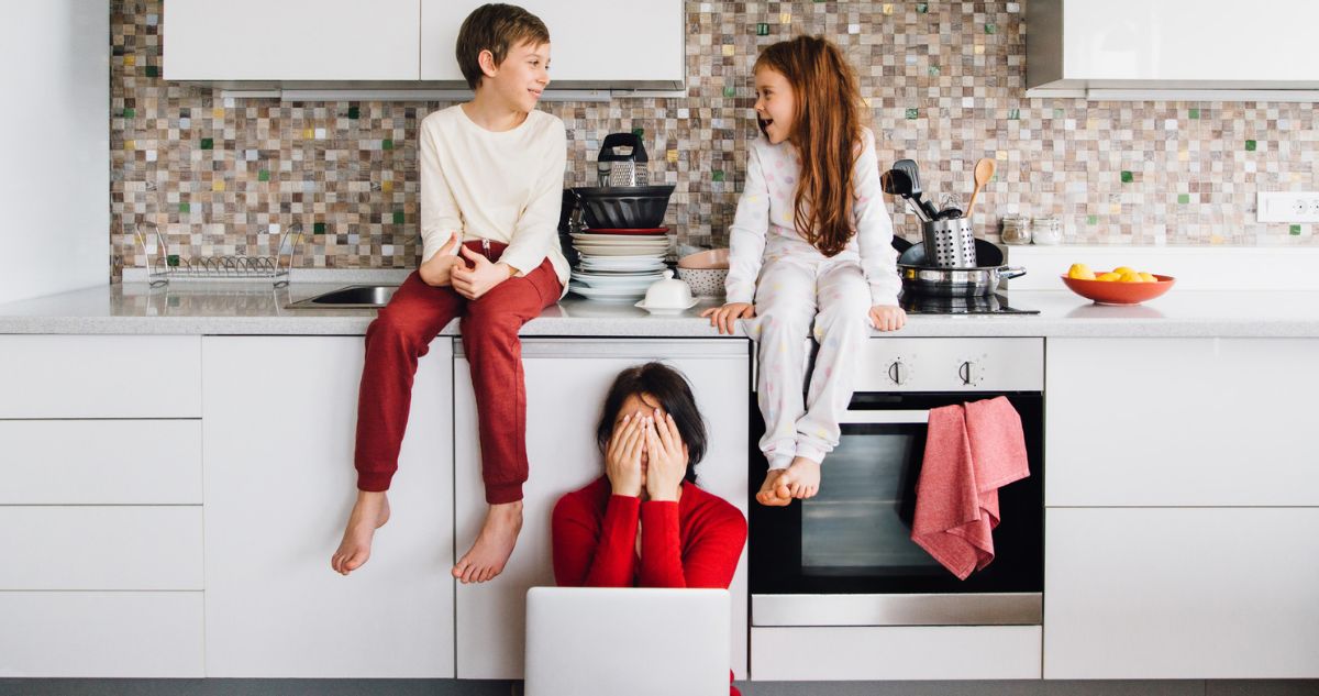anya fáradtan a konyhában a földön ül ölében laptoppal, gyerekei a pulton ülnek beszélgetve