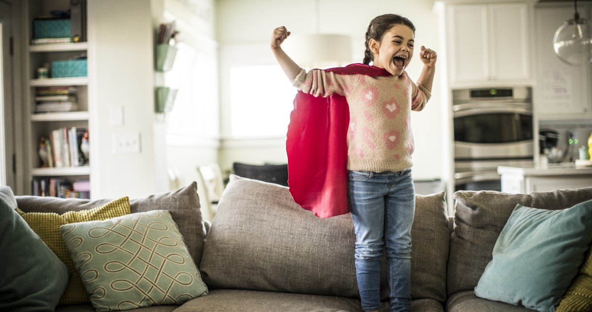 kislány szuperhősnek öltözve áll a kanapén