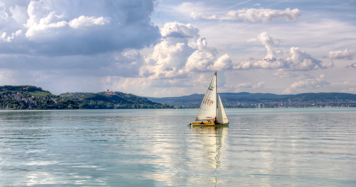 egy vitorlás úszik a kékes vízű Balatonon, a háttérben hegyek