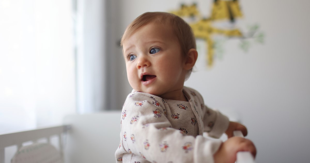 képen egy nagyjából egyéves kislány baba látható, aki a kiságyában áll, fehér, virágmintás ruhában