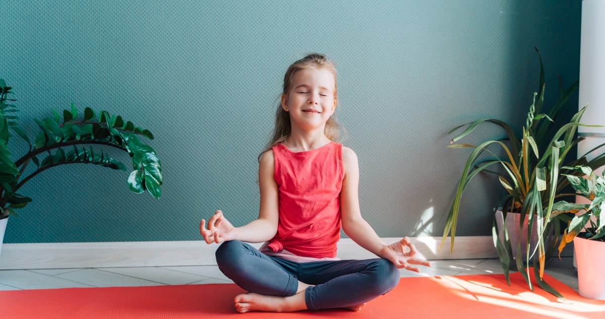 kisgyerek-legzes - Gyerekszoba A képen egy narancssárga jógamatracon ül egy kisiskolás lány, piros felsőben és fekete nadrágban, meditálós ülésben, és mosolyogva, csukott szemmel lélegzik be. A háttérben szobanövények.
