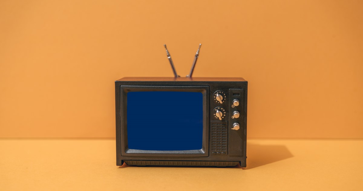 A képen narancssárga háttér előtt egy régi, barna, antennás, tekerő-kapcsolós televízió-készülék látható.