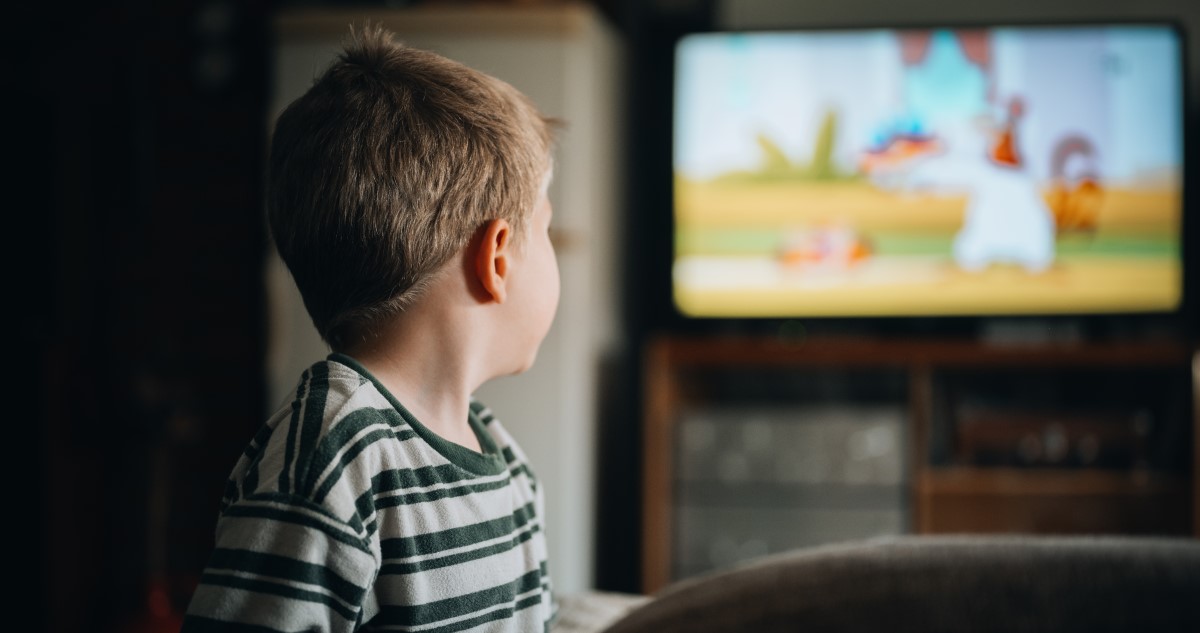 A képen egy kisfiú látható, aki a tévétől távolabb állva nézi a képernyőt, amin egy mesefilm megy.