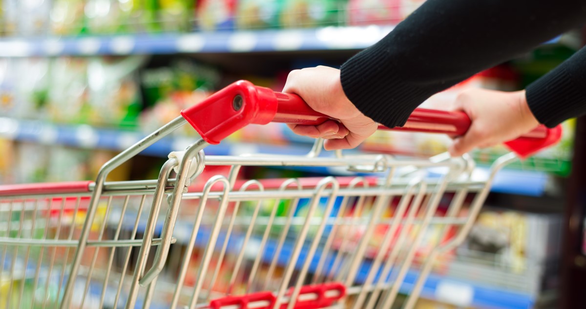 A fényképen közelről látható egy nő keze, aki egy bevásárló kocsit tol egy üzletben, a háttérben pedig elmosódottan a bolt polcai a termékekkel.