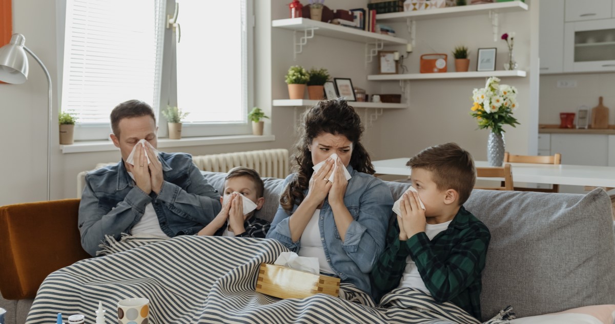 A képen egy család látható, a nappaliban ülnek, pokróccal betakarva, mindenki orrot fúj, a szülők és a két kisgyerekük is.