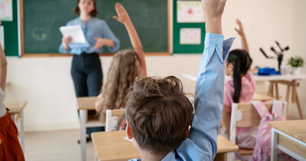 A képen egy tanteremben láthatunk kisiskolásokat. Köelről egy kék inges kisfiú hátát, aki jelentkezik, előtte, több jelentkező kislány. A háttérben a tábla előtt elmosódottan látható, hogy egy tanárnő áll.