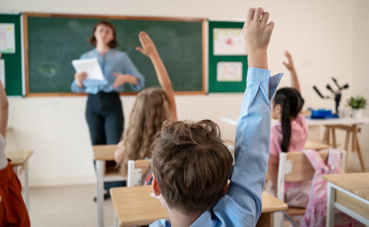 A képen egy tanteremben láthatunk kisiskolásokat. Köelről egy kék inges kisfiú hátát, aki jelentkezik, előtte, több jelentkező kislány. A háttérben a tábla előtt elmosódottan látható, hogy egy tanárnő áll.