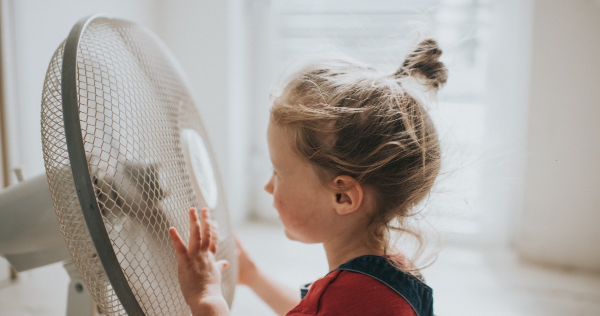 A képen oldalról látható egy kislány, aki a működő ventillátor előtt áll.
