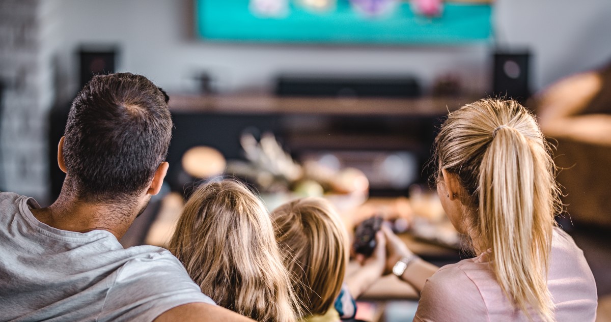 A képen egy négytagú család látható hátulról, akik a nappaliban ülnek és tévét néznek.