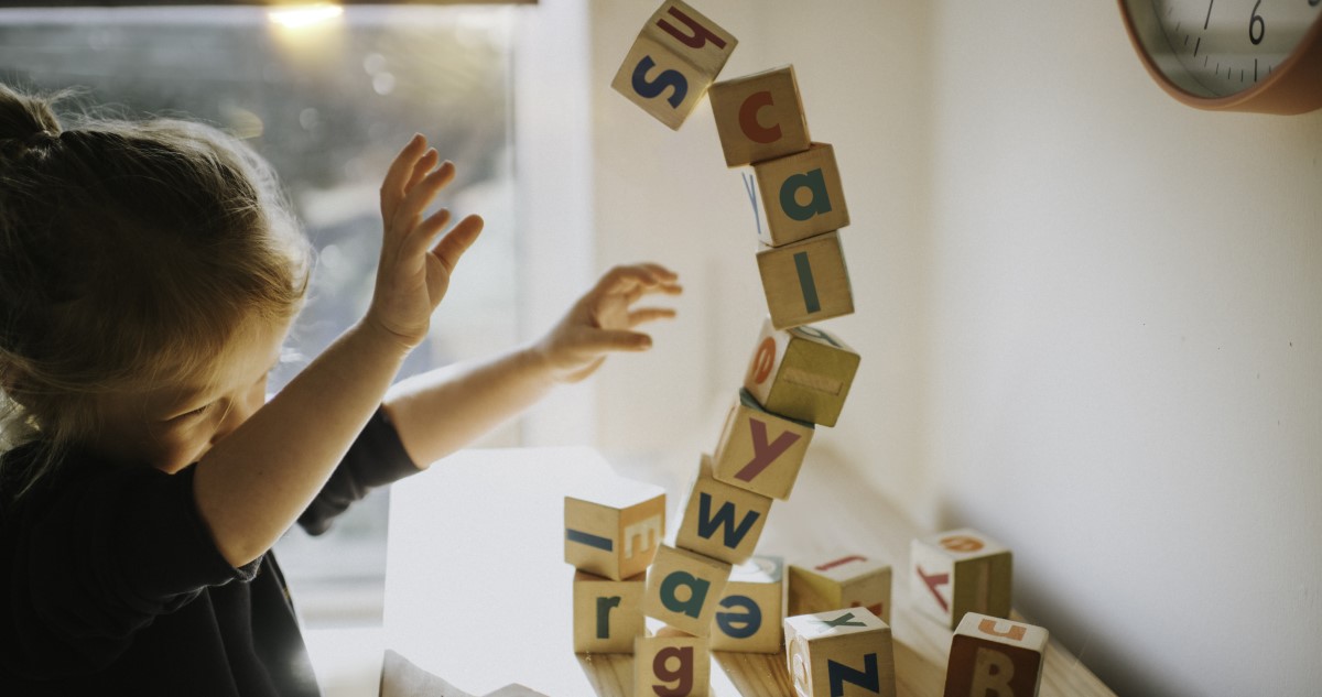 A képen egy kislány látható, aki betűs építőkockákból épült tornyot borít le.