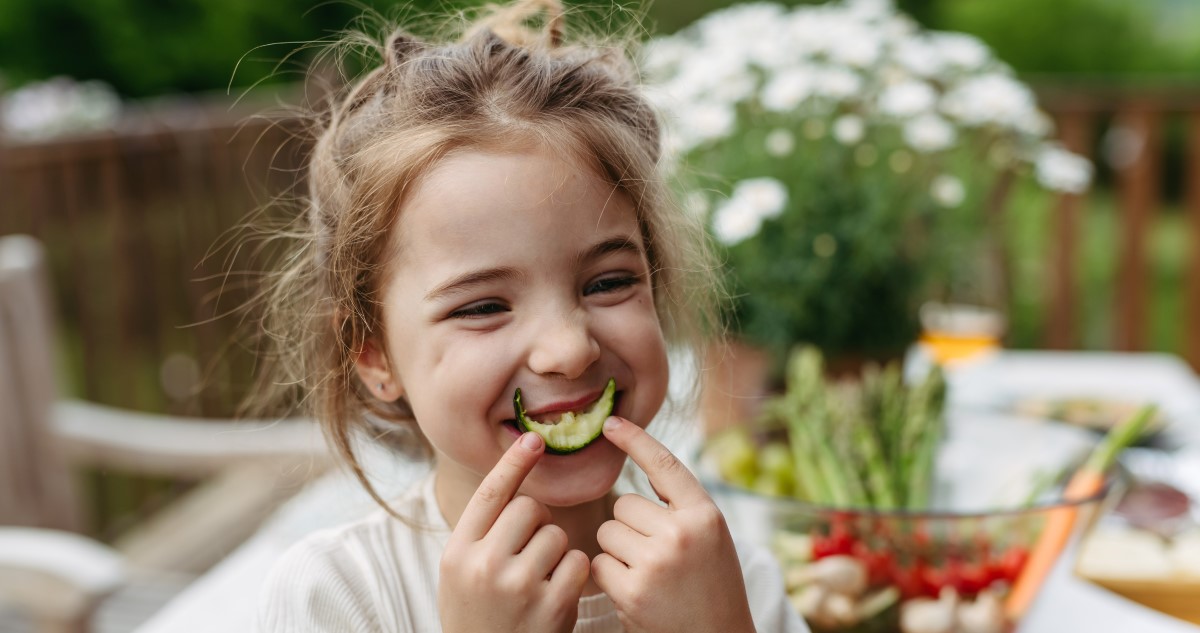 kislany-zoldseget-eszik - Gyerekszoba A képen egy kislány látható, aki egy fél karika uborkát tart a szája elé, mintha egy mosoly lenne.