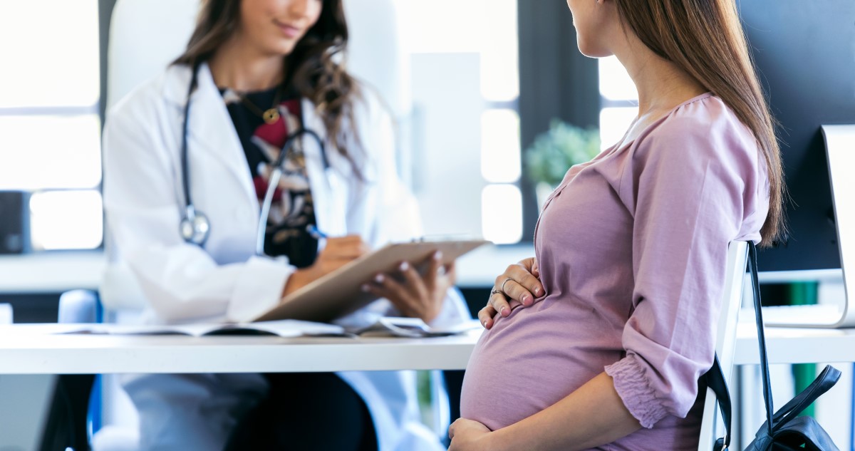 A képen egy kismama látható, aki várandósgondozáson vesz részt, az orvossal beszél a rendelőben.