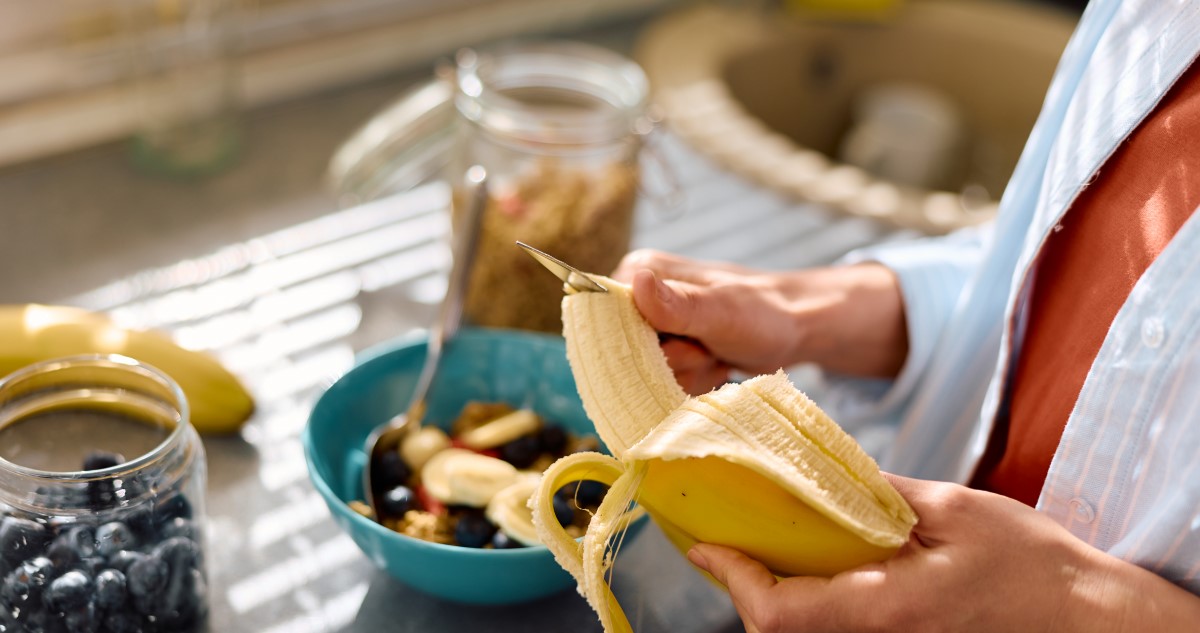 banan-gyumolcstal - Gyerekszoba A képen banánt karikáz egy nő, akinek csak a keze látszik. A háttérben egy tálban gyümölcsök látszanak, egy üvegben pedig áfonya.