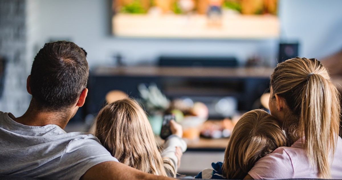 A képen egy négytagú család látható, akik a nappaliban a kanapén ülve néznek egy mesét a tévében.