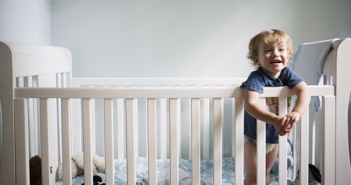 A képen egy kisgyerek látható, aki a rácsos kiságyban áll a matracon.