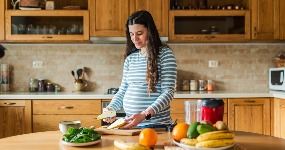 A képen egy kismama látható, aki körtét vág fel, és gyümölcsökből, salátából smoothiet készít.