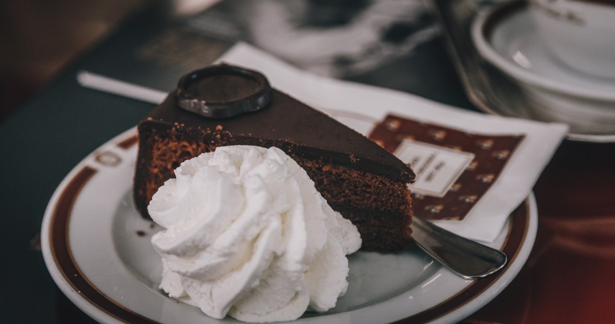 A képen egy szelet klasszikus Sacher-torta látható, egy adag tejszínhabbal, a háttérben kávé.